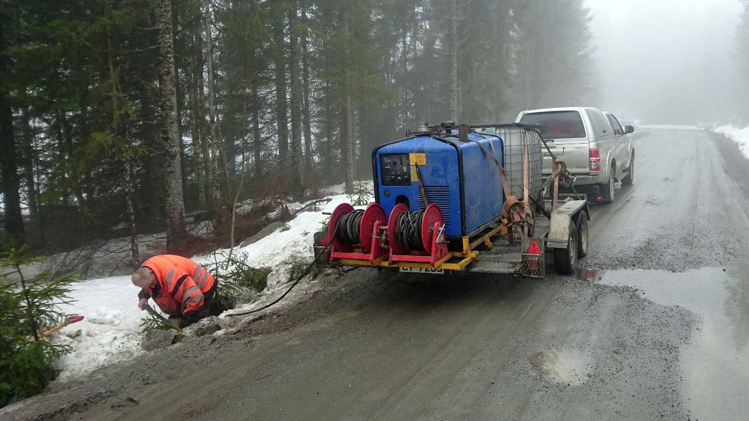 Bil med henger som står på en skogsbilveg. På hengeren står et tineapperat. En mann står i grøfta og jobber med å tine opp en stikkrenne. Foto. står et